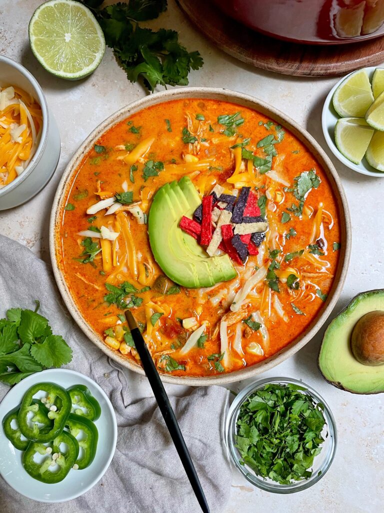 Chicken enchilada soup in a bowl with toppings and garnishes around the bowl.