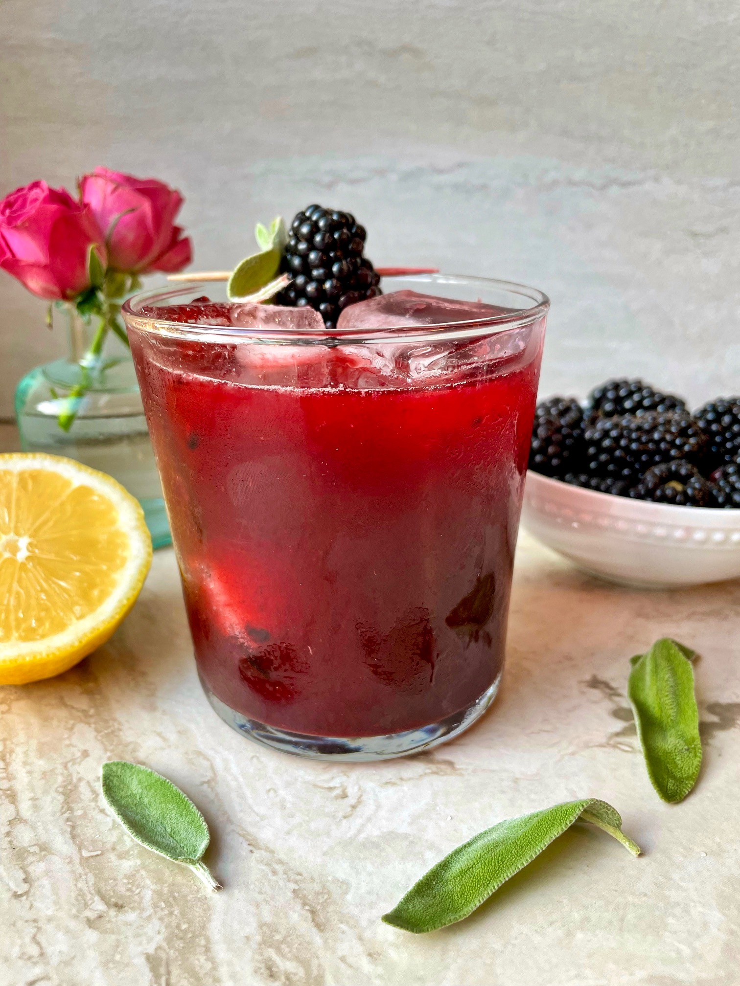a side shot of the completed margarita with a lemon half, sage leaves, flowers and blackberries in a bowl surrounding the drink