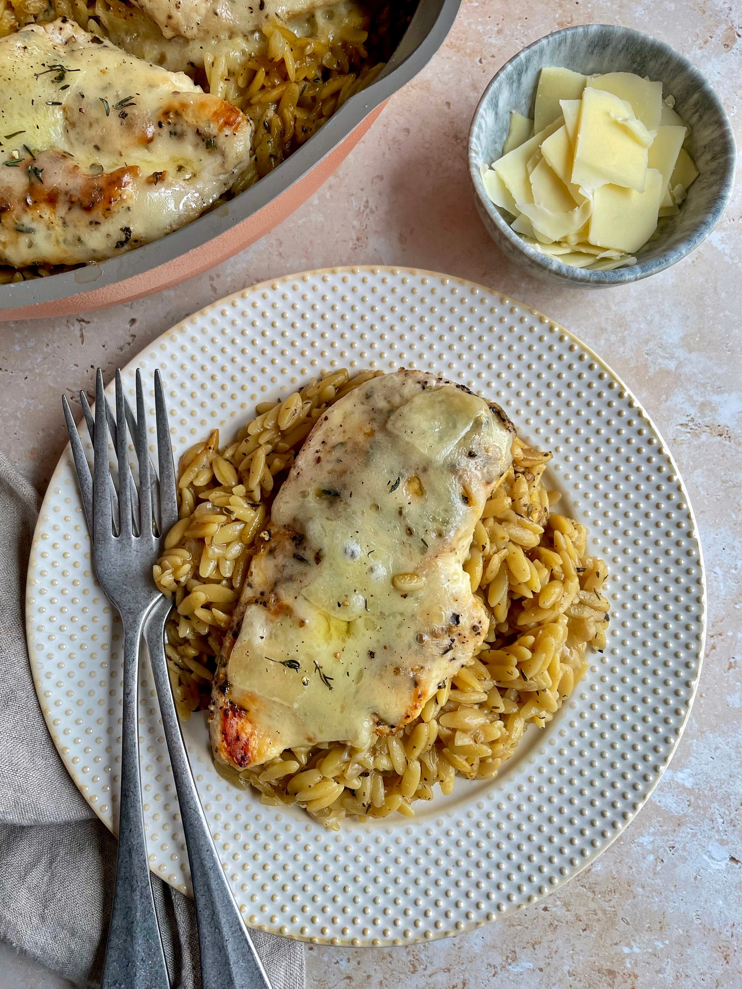 A plate of french onion chicken and orzo with the pan and a bowl of shaved gruyere next to it.