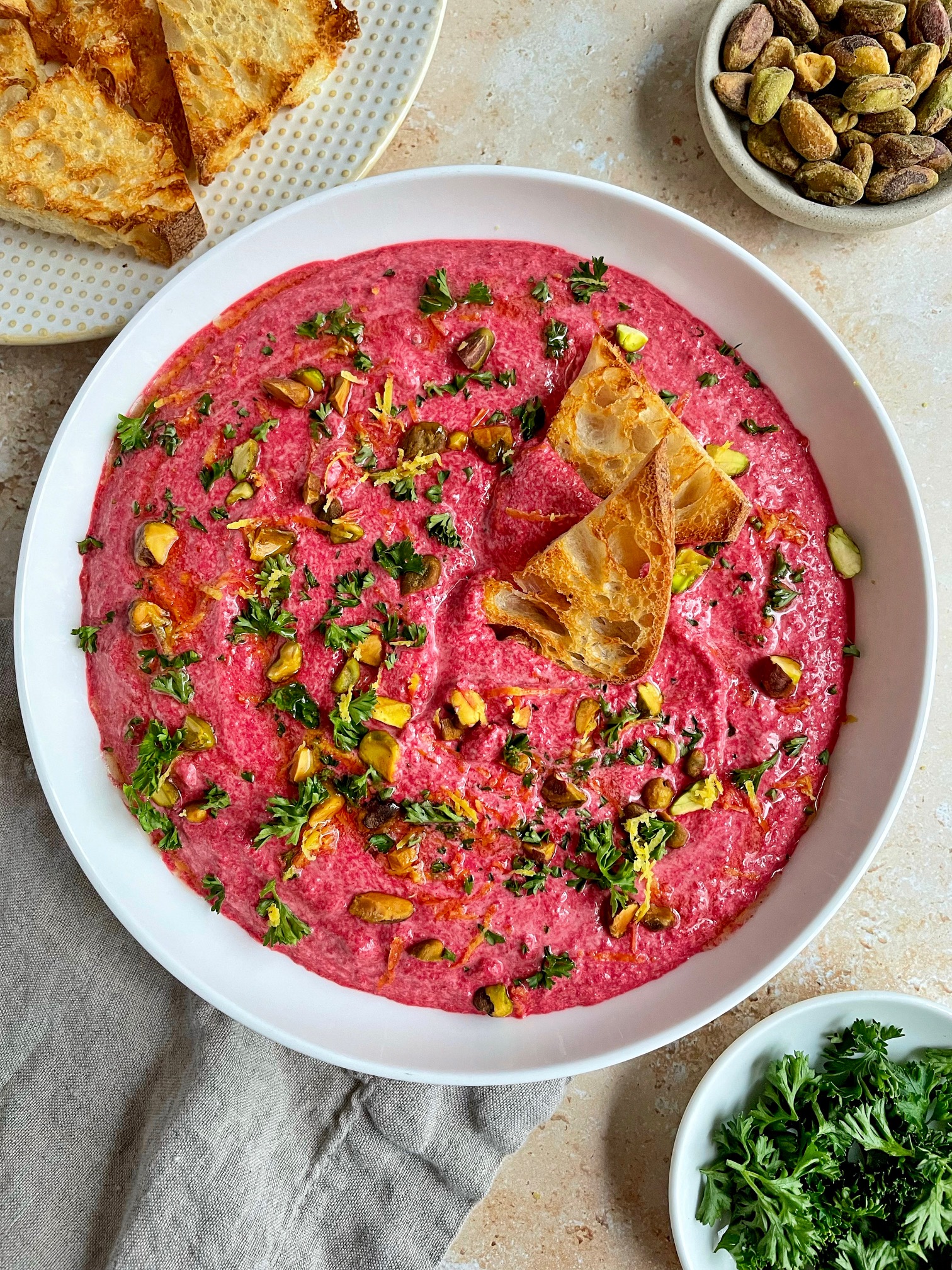 a bowl of beet and goat cheese dip with two pieces of toast in the tip and garnishes around the bowl.