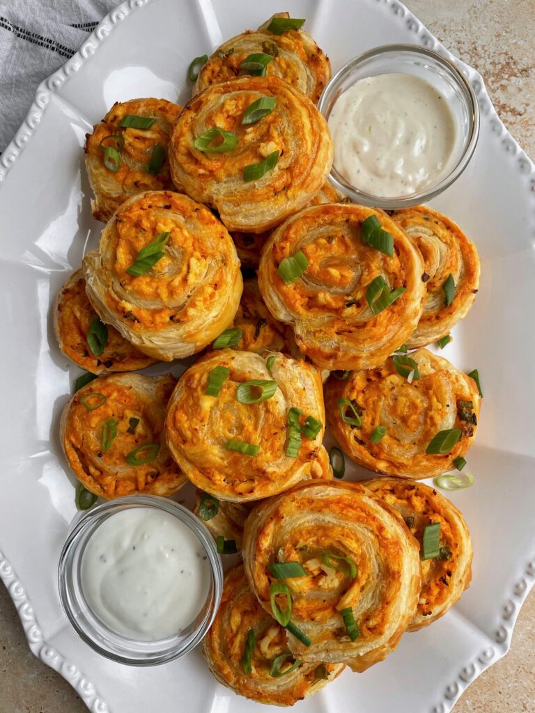 A platter of buffalo chicken pinwheels garnished with scallions and served with blue cheese and ranch.