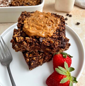 Two pieces of chocolate baked oatmeal on a plate with a fork, strawberries and peanut butter topping.