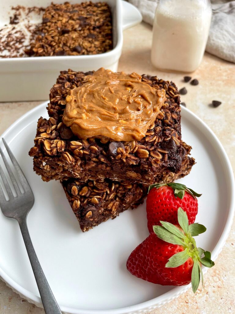 Two pieces of chocolate baked oatmeal on a plate with a fork, strawberries and peanut butter topping.