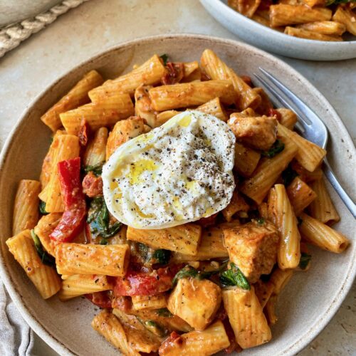 A bowl of creamy tomato pesto pasta with chicken topped with half a ball of burrata.