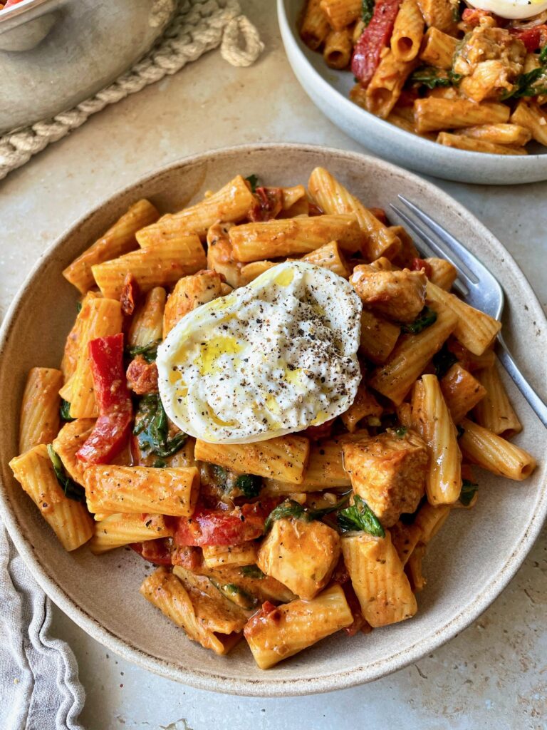A bowl of creamy tomato pesto pasta with chicken topped with half a ball of burrata.