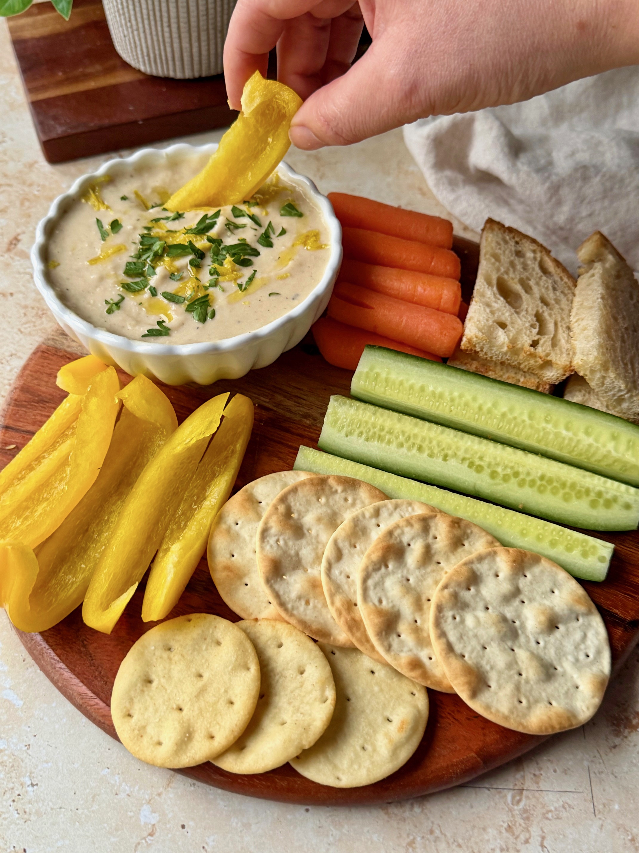 Dipping a bell pepper into the dip that is on a wooden board with fresh veggies, toasted bread and crackers.